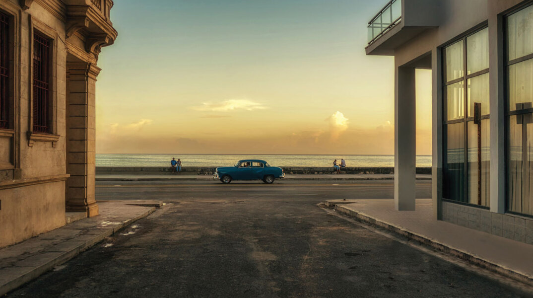 Ένα μπλε αυτοκίνητο φαίνεται στο βάθος της φωτογραφίας, ανάμεσα σε δύο κτήρια και με τη θάλασσα στο φοντο, ώρα ηλιοβασιλέματος