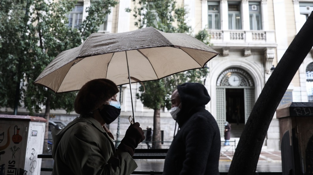 Γυναίκα περπατά κρατώντας την ομπρέλα της - Στο βάθος άνδρας έχει φορέσει την κουκούλα για να προστατευτεί από τη βροχή