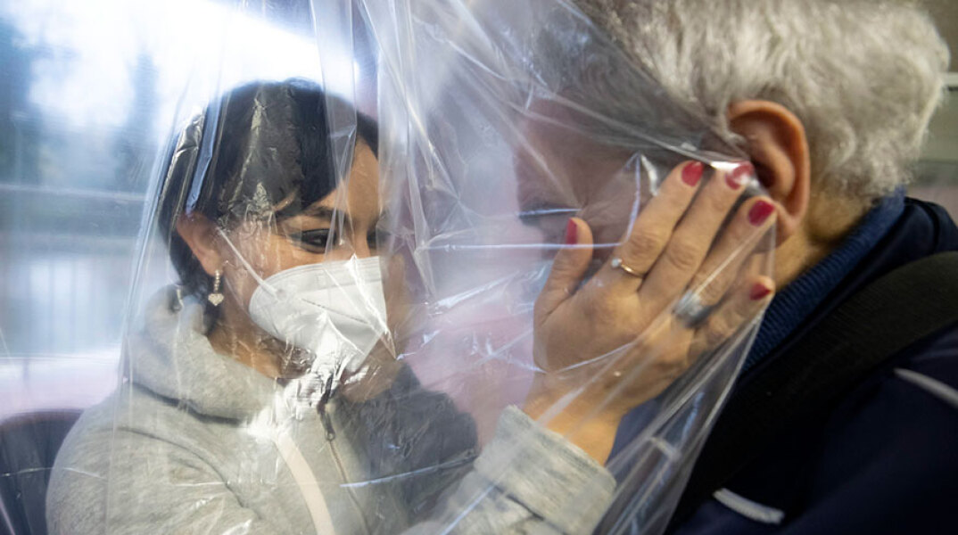 Γυναίκα με μάσκα στην Ιταλία αγκαλιάζει ηλικιωμένο σε γηροκομείο, έχοντας ανάμεσά τους μια ειδική πλαστική κουρτίνα