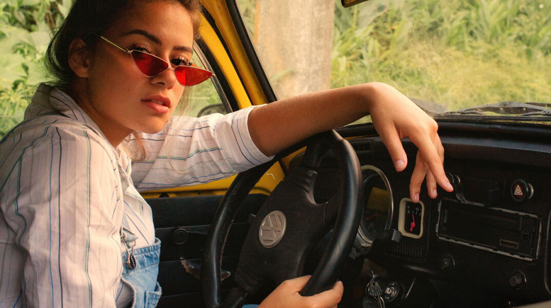 Γυναίκα καθισμένη σε αυτοκίνητο, κρατάει τιμόνι και κοιτάζει την κάμερα