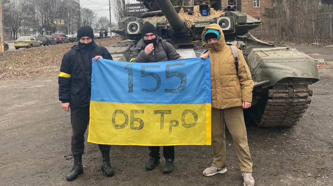 Τρεις νεαροί άνδρες κρατούν σημαία της Ουκρανίας μπροστά από τανκ