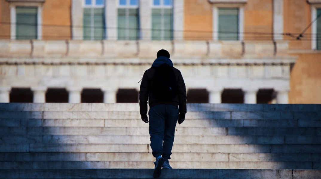 Νεαρός ανεβαίνει τα σκαλιά της πλατείας Συντάγματος έχοντας μπροστά του τη Βουλή των Ελλήνων