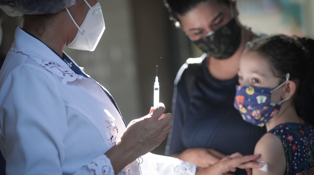 Παιδί στην αγκαλιά του γονέα του κοιτά το εμβόλιο κατά του κορωνοϊού λίγο πριν το κάνει η νοσηλεύτρια