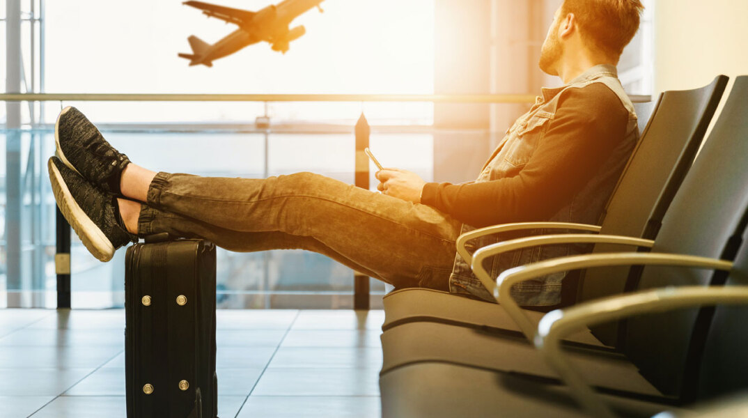 Άνδρας σε κάθισμα αεροδρομίου με ταπόδια απλωμένα πάνω στη βαλίτσα κοιτάζει έξω από την τζαμαρία τα αεροπλάνα