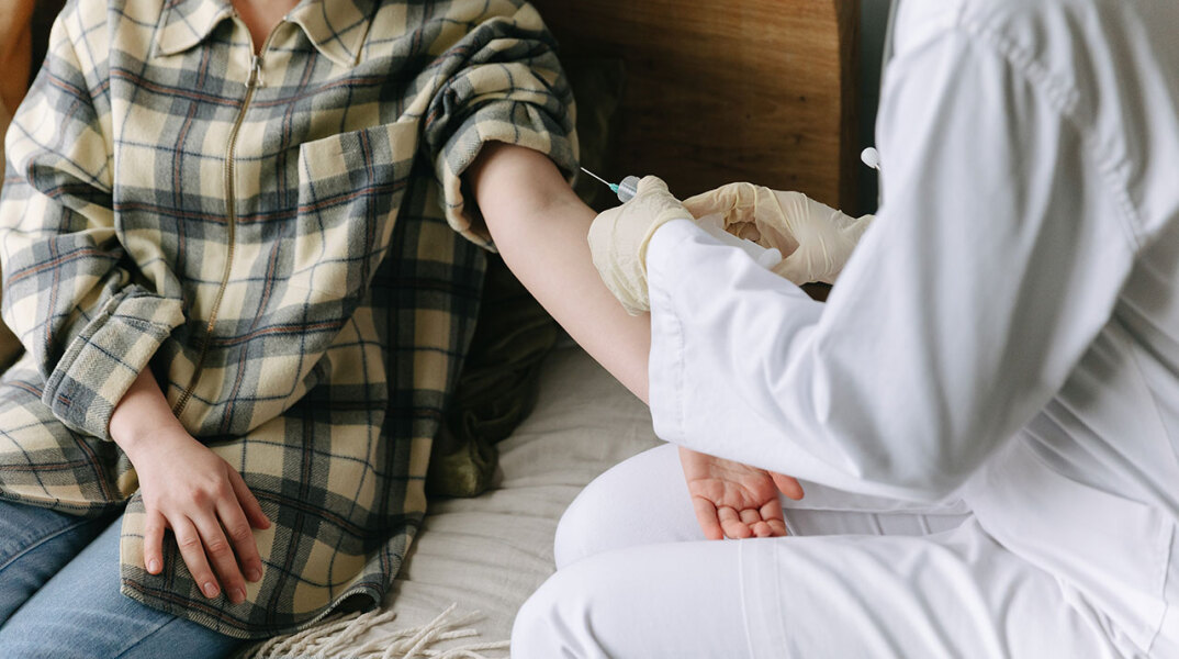 Γυναίκα με καρό πουκάμισο και απλωμένο χέρι, δίπλα της νοσηλευτής με ένεση