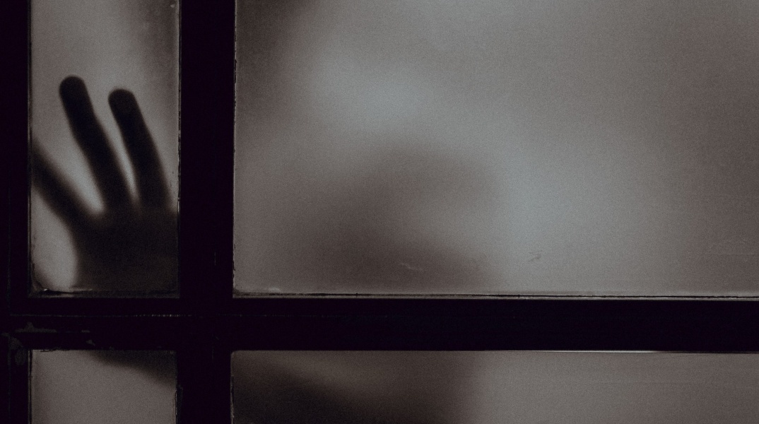 Χέρι και πρόσωπο διακρίνεται πίσω από κλειστή πόρτα σε ημίφως