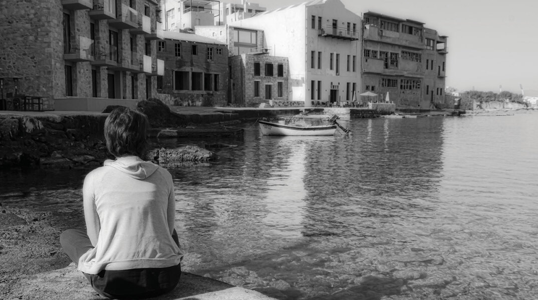 Ασπρόμαυρη φωτογραφία γυναίκας καθισμένης με γυρισμένη την πλάτη και φόντο τη θάλασσα