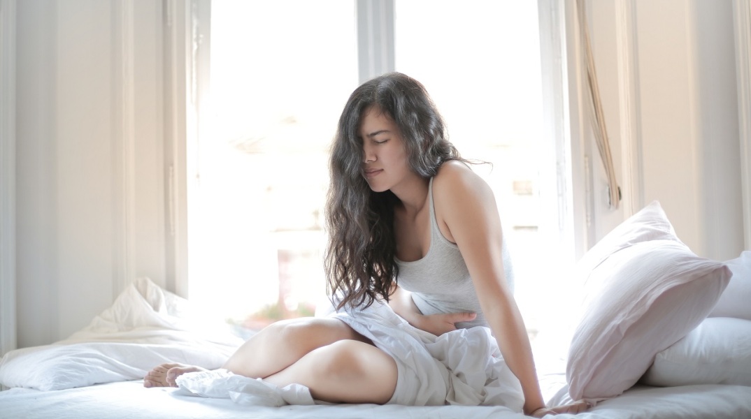 Γυναίκα καθισμένη σε κρεβάτι κρατάει την κοιλιά της με δυσφορία