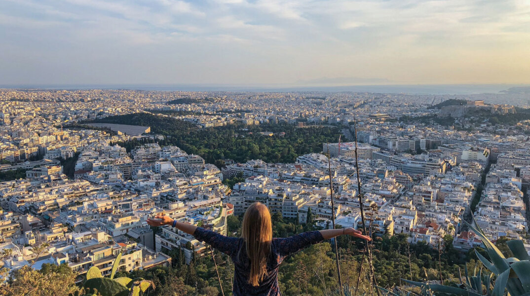 Κοπέλα στέκεται όρθια με γυρισμένη πλάτη με φόντο την Αθήνα από ψηλά