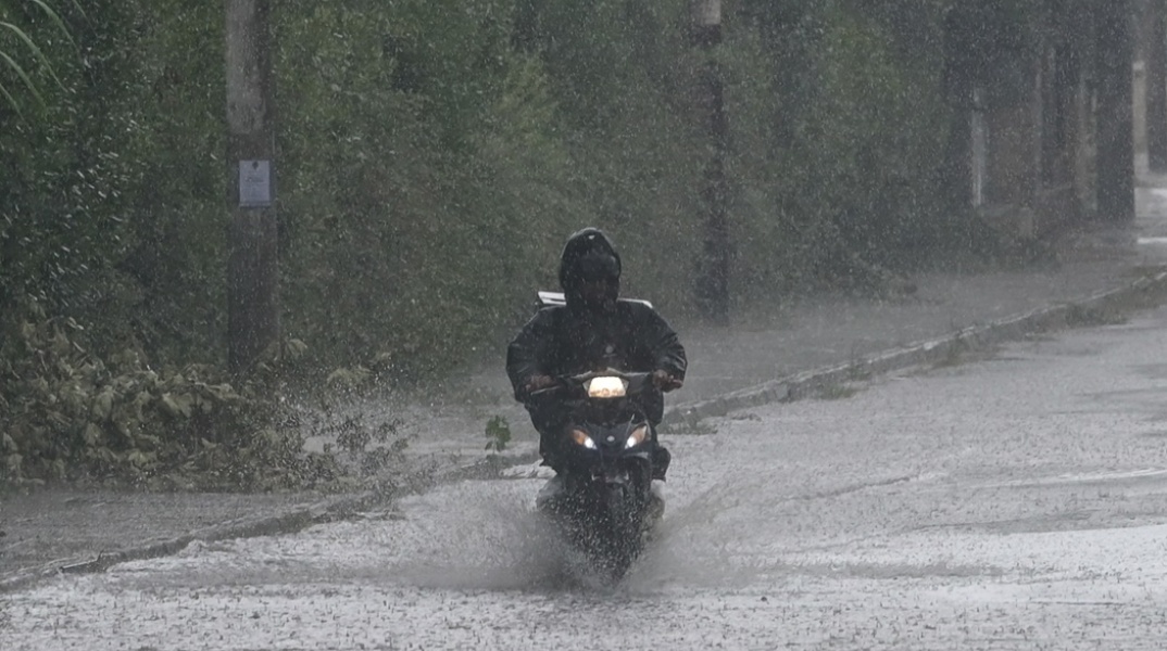 Κακοκαιρία: Διανομέας με αδιάβροχο οδηγεί μηχανάκι υπό καταρρακτώδη βροχή σε πλημμυρισμένο δρόμο