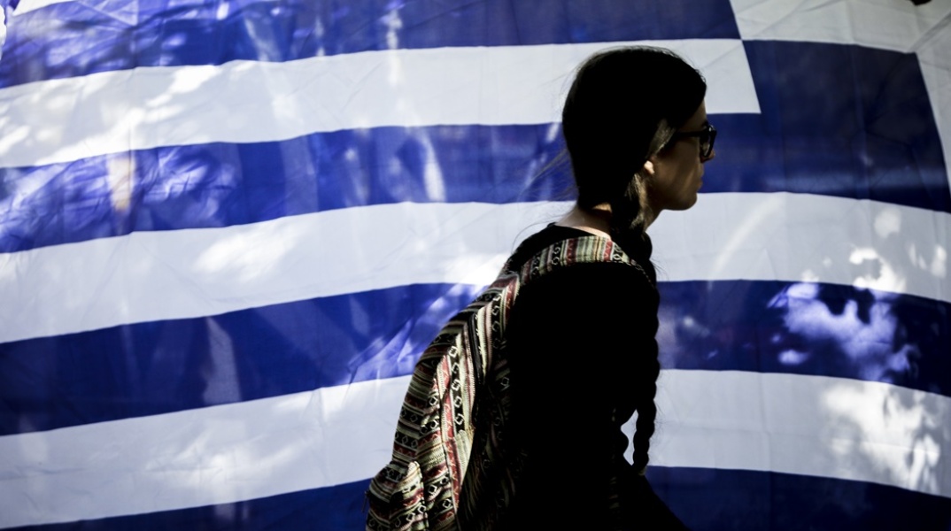 Γυναίκα προχωρά μπροστά από ελληνική σημαία που κυματίζει