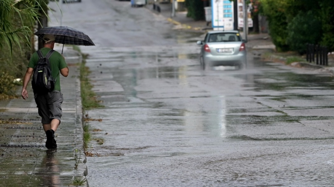 Πολίτης με ομπρέλα σε πεζοδρόμιο και δίπλα του αυτοκίνητο διασχίζει πλημμυρισμένο δρόμο