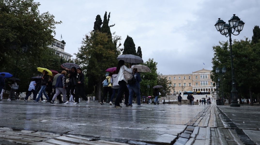 Πολίτες στο κέντρο της Αθήνας κρατούν ομπρέλες διασχίζοντας τον δρόμο που οδηγεί στην πλατεία Συντάγματος