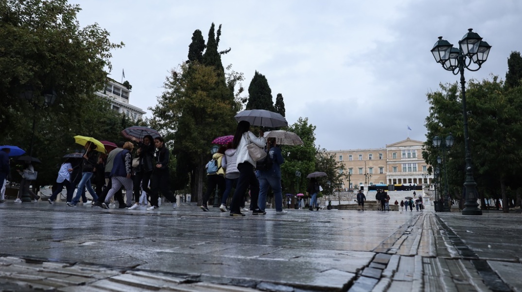 Πολίτες στο κέντρο της Αθήνας κρατούν ομπρέλες διασχίζοντας τον δρόμο που οδηγεί στην πλατεία Συντάγματος