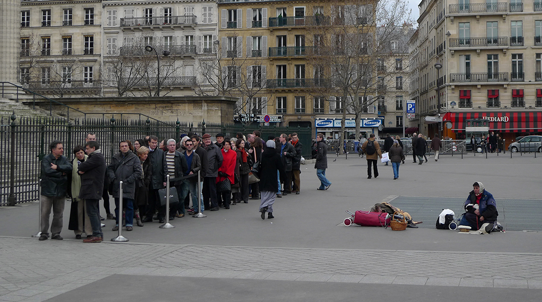Άνθρωποι που περιμένουν σε μια ουρά και μια άστεγη γυναίκα κάθεται κοντά τους στην πλατεία Madeleine στο Παρίσι