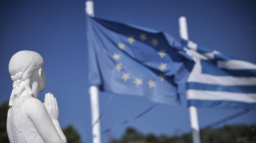 Άγαλμα με χέρια σε στάση προσοχής που κοιτά την ελληνική σημαία και τη σημαία της Ευρωπαϊκής Ένωσης