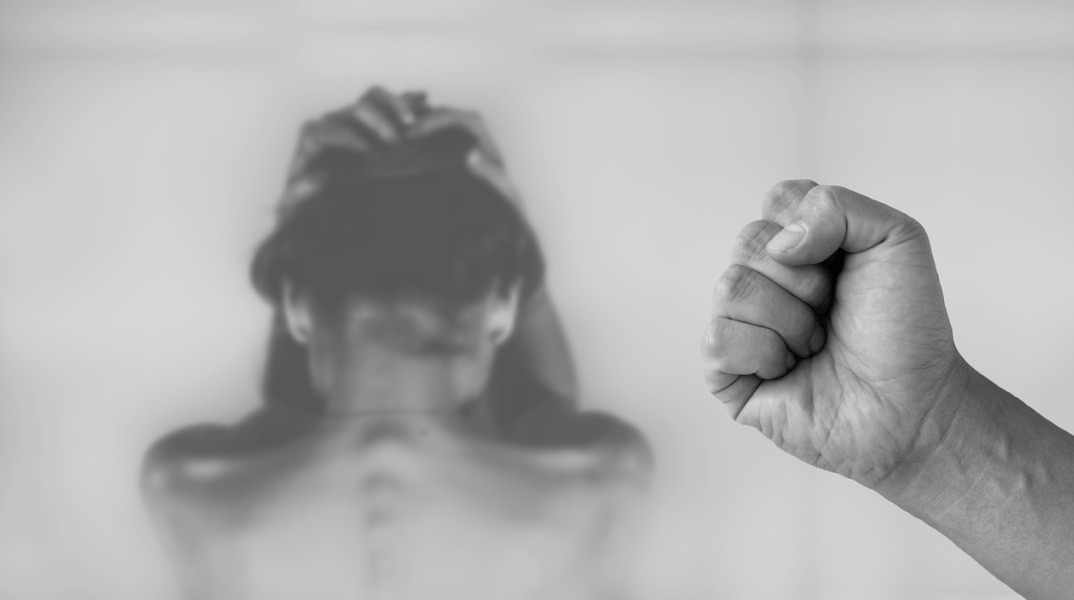 Γυναίκα θύμα ενδοοικογενειακής βίας γυρισμένη πλάτη, όσο μία γροθιά φαίνεται στα δεξιά της φωτογραφίας