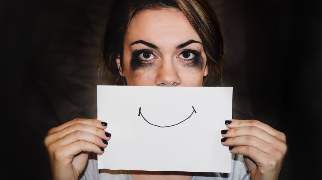 Γυναίκα με κλαμένα μάτια κρύβει τα χείλη της με ένα χαρτί που έχει πάνω του ζωγραφισμένο χαμόγελο