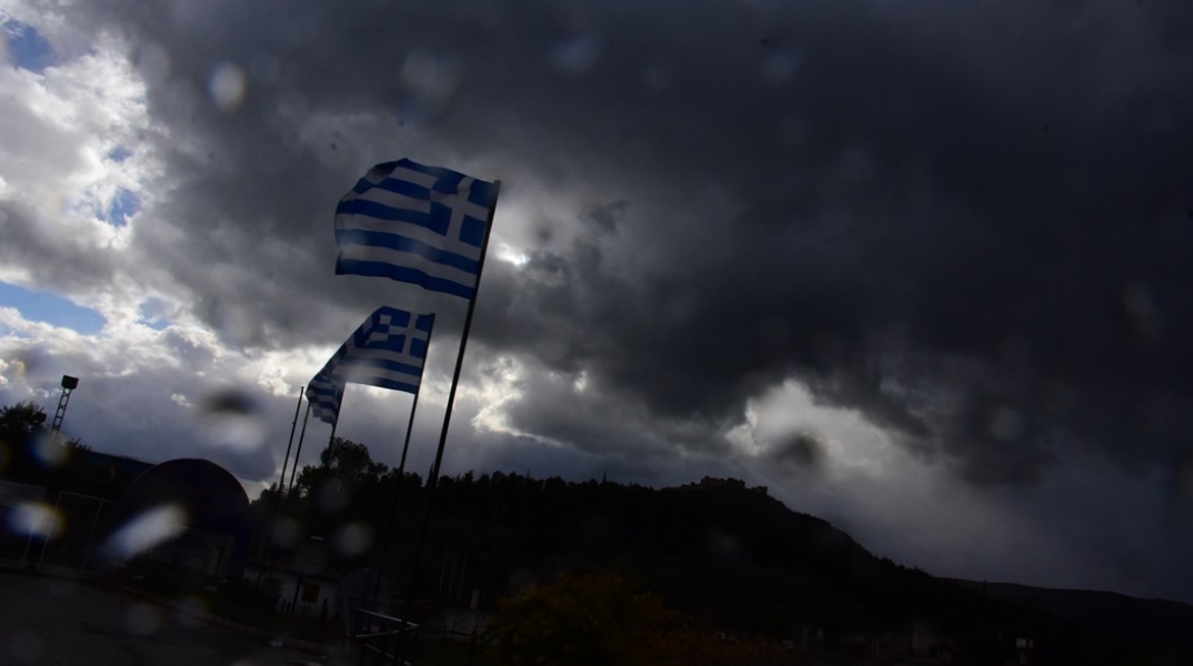 Σταγόνες βροχής πάνω σε τζάμι και στο φόντο ελληνικές σημαίες 