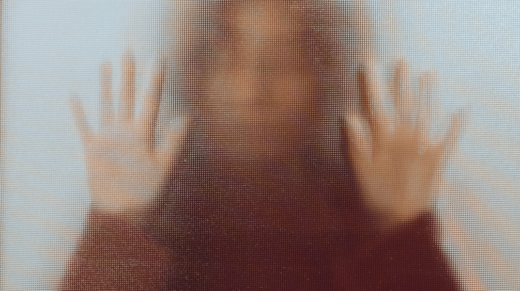 Γυναίκα πίσω από ημιδιάφανο τζάμι με κολλημένα τα χέρια της στην επιφάνεια σε στάση που παραπέμπει σε φόβο και απόγνωση