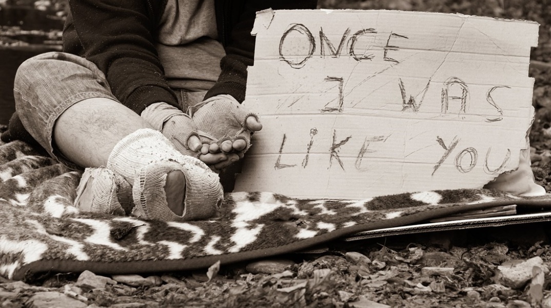 Άστεγος με τρύπια και βρώμικα ρούχα με χαρτόνι στο οποίο γράφει «Κάποτε ήμουν σαν εσένα»