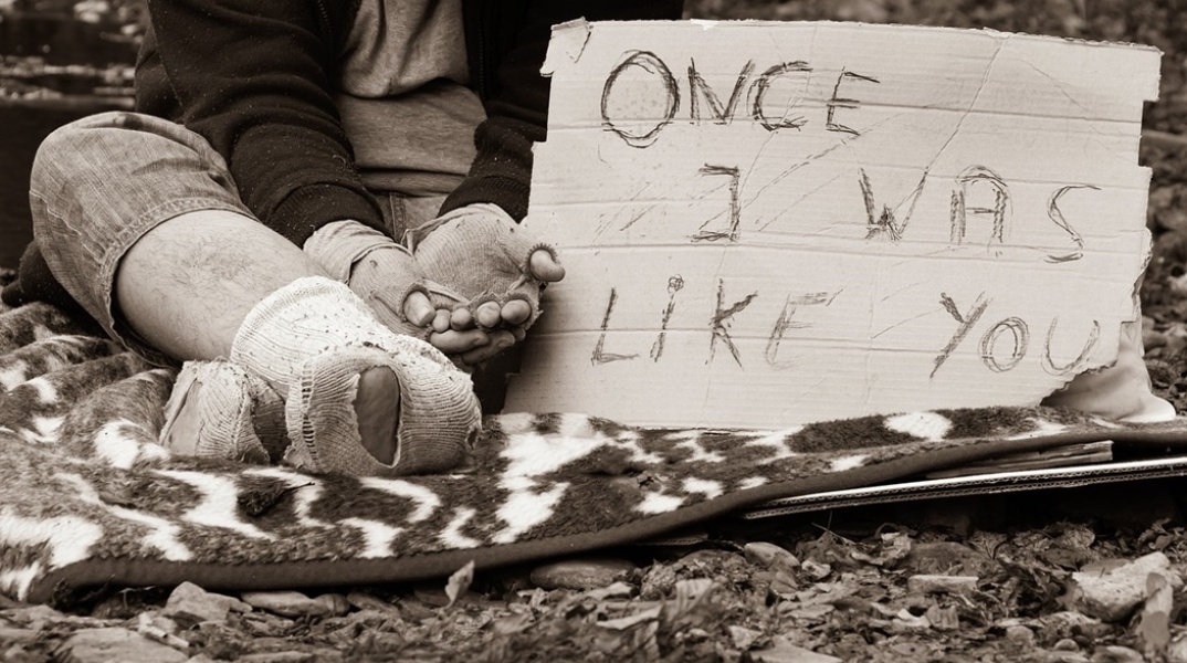 Άστεγος με τρύπια και βρώμικα ρούχα με χαρτόνι στο οποίο γράφει «Κάποτε ήμουν σαν εσένα»