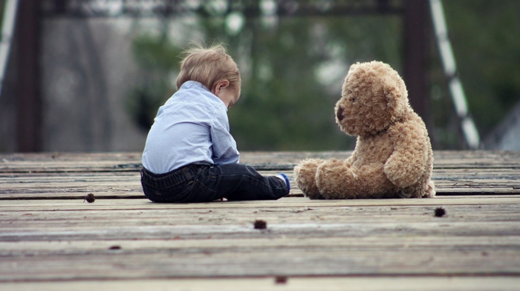 Παιδί κάθεται σε ξύλινο πάτωμα μαζί με το αρκουδάκι του