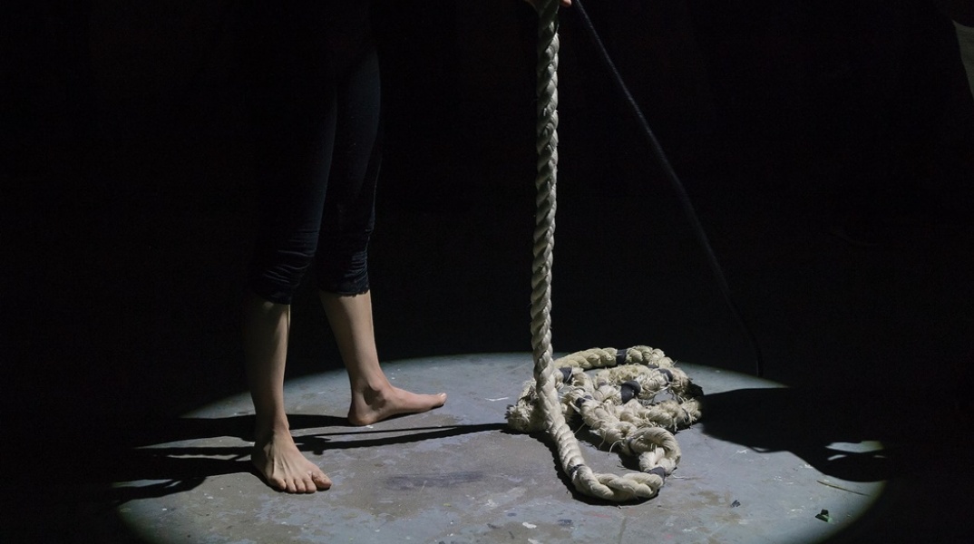 Άτομο με γυμνά πόδια κρατά χοντρό σκοινί σε σκοτεινό δωμάτιο - Εικόνα που παραπέμπει σε αυτοκτονία δι' απαγχονισμού