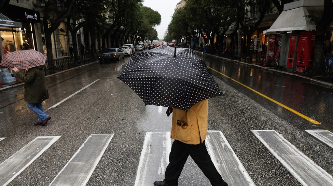 Πολίτες περνούν διάβαση κρατώντας ομπρέλα για την προστασία τους από βροχόπτωση