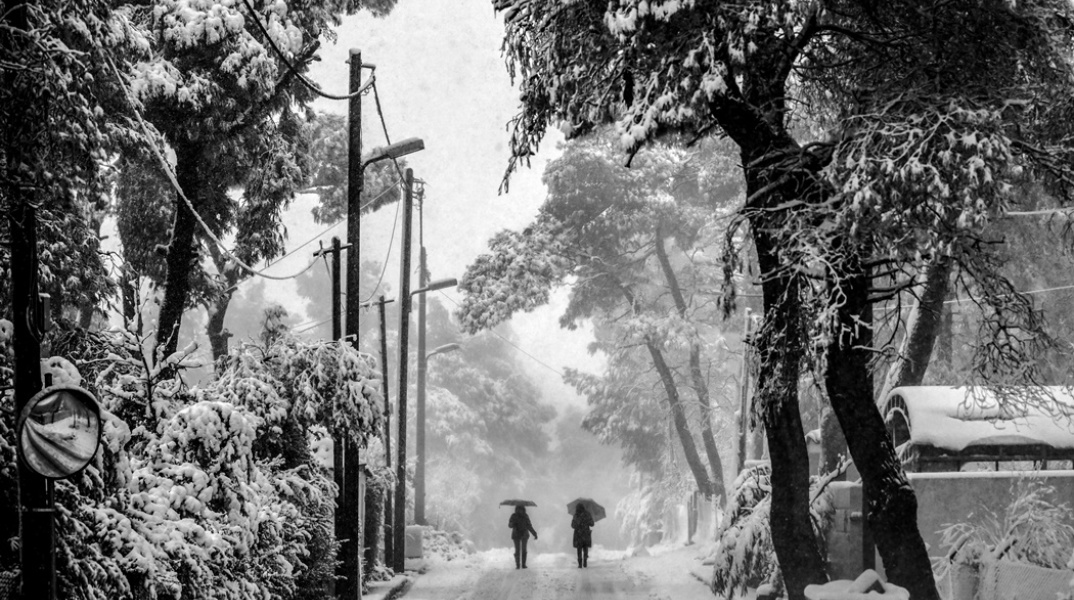 Χιονισμένο τοπίο και δύο πολίτες περπατούν κρατώντας την ομπρέλα τους