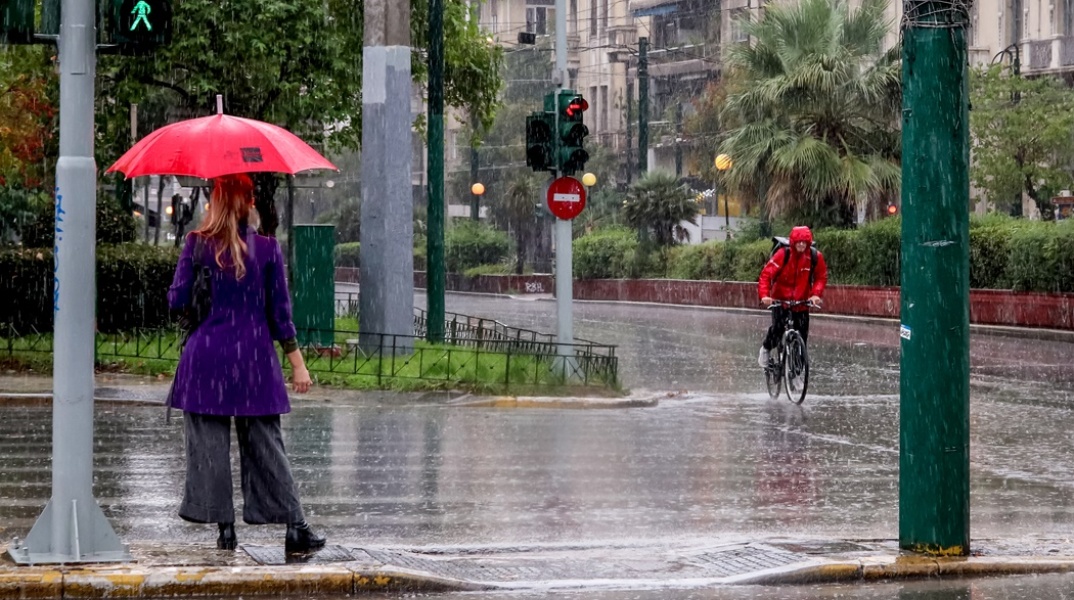 Γυναίκα με ομπρέλα περιμένει σε διάβαση πεζών την ώρα που ποδηλάτης με αδιάβροχο κινείται στους δρόμους της Αθήνας