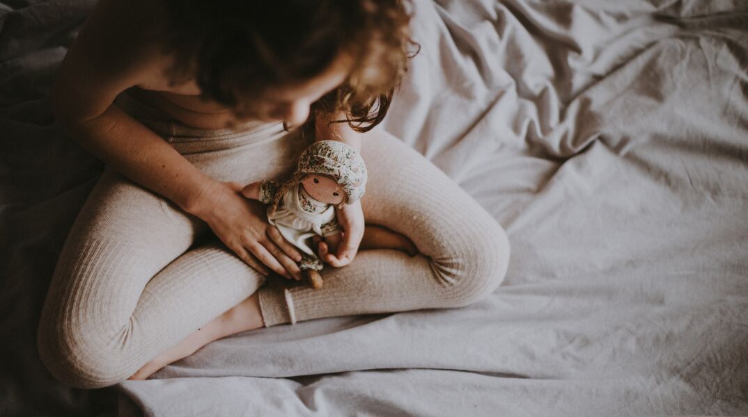 Παιδί κάθεται οκλαδόν σε κρεβάτι και κρατά στα χέρια του κούκλα