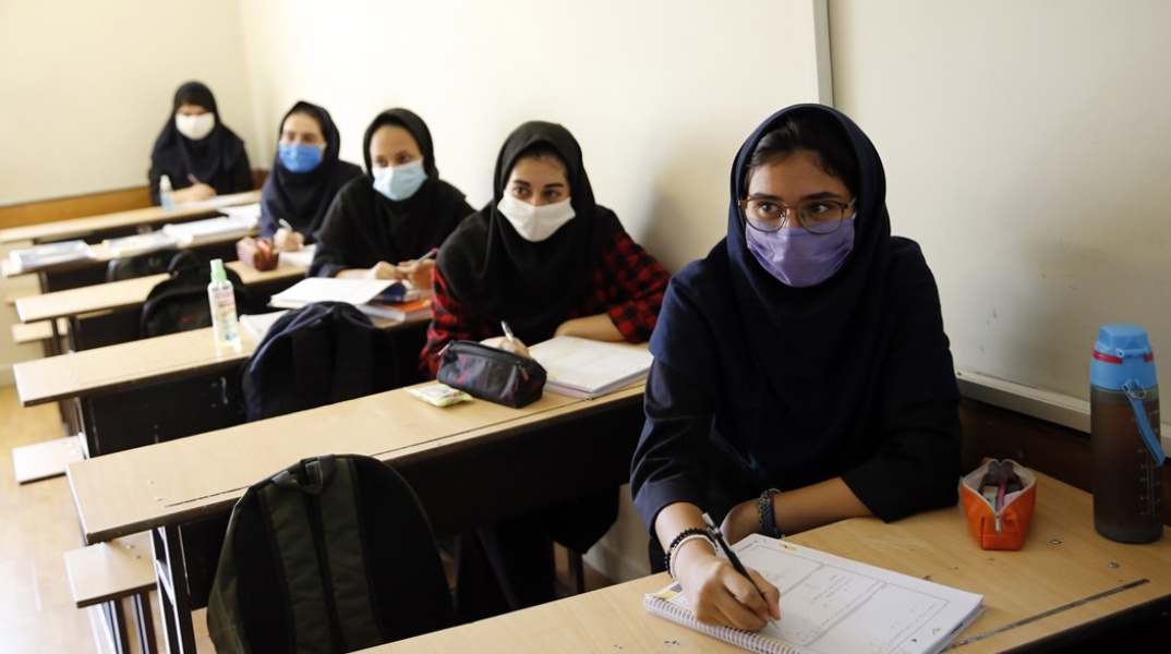 Μαθήτριες σε σχολείο στο Ιράν κάθονται στα έδρανα έχοντας ανοίξει τα τετράδια τους και φορώντας τις μάσκες τους