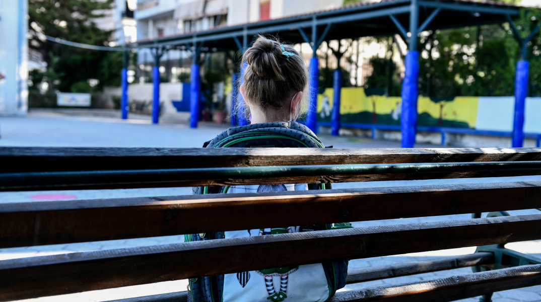 Μαθήτρια Δημοτικού με την τσάντα τους ώμους κάθεται σε παγκάκι στο προαύλιο του σχολείου