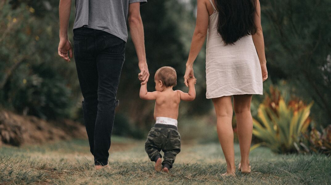 Πατέρας και μητέρα κρατούν το μωρό τους και περπατάνε όλοι μαζί, ξυπόλυτοι στη φύση