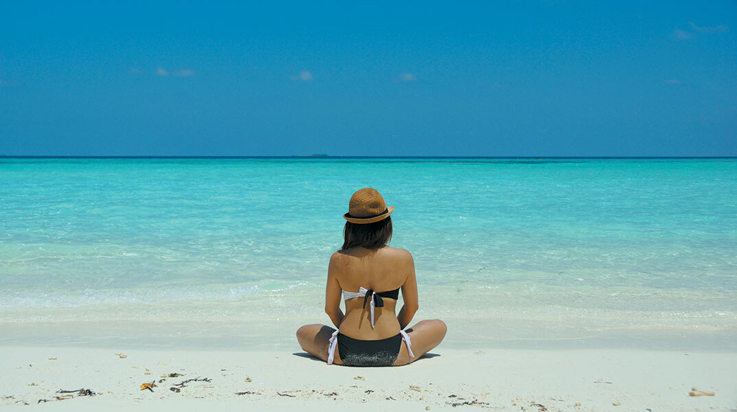 Κοπέλα με μαγιό κάθεται σε αμμώδη παραλία