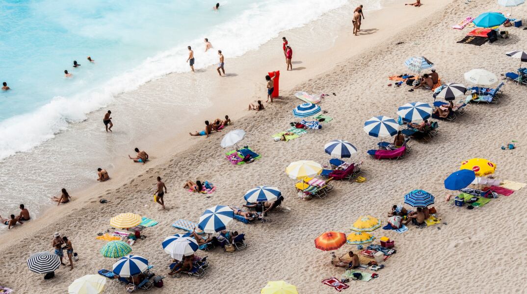 Λουόμενοι απολαμβάνουν τον ήλιο, το κολύμπι και τα παιχνίδια σε παραλία της Λευκάδας - Στημένες ομπρέλες και απλωμένες πετσέτες στην αμμουδιά