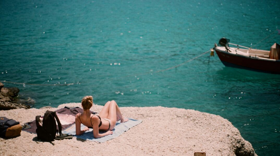 Γυναίκα με την πετσέτα της απολαμβάνει την ηλιοθεραπεία δίπλα στη θάλασσα μετά το μπάνιο της