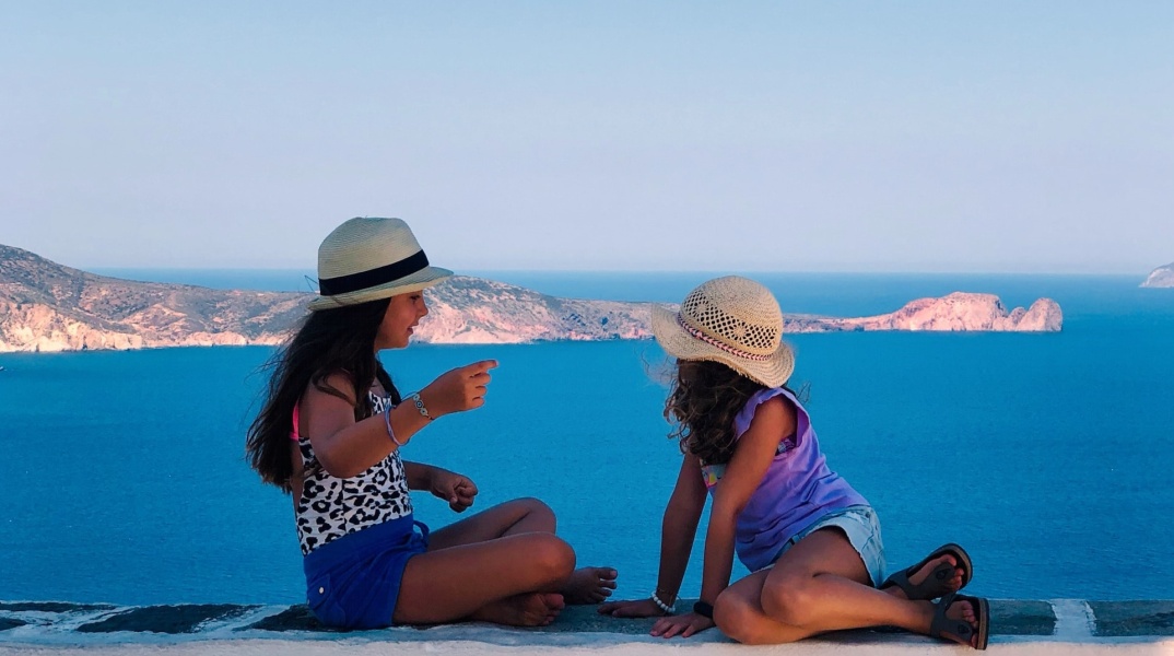 Παιδιά κάθονται σε πεζουλάκι νησιού και φωτογραφίζονται με φόντο τη θέα