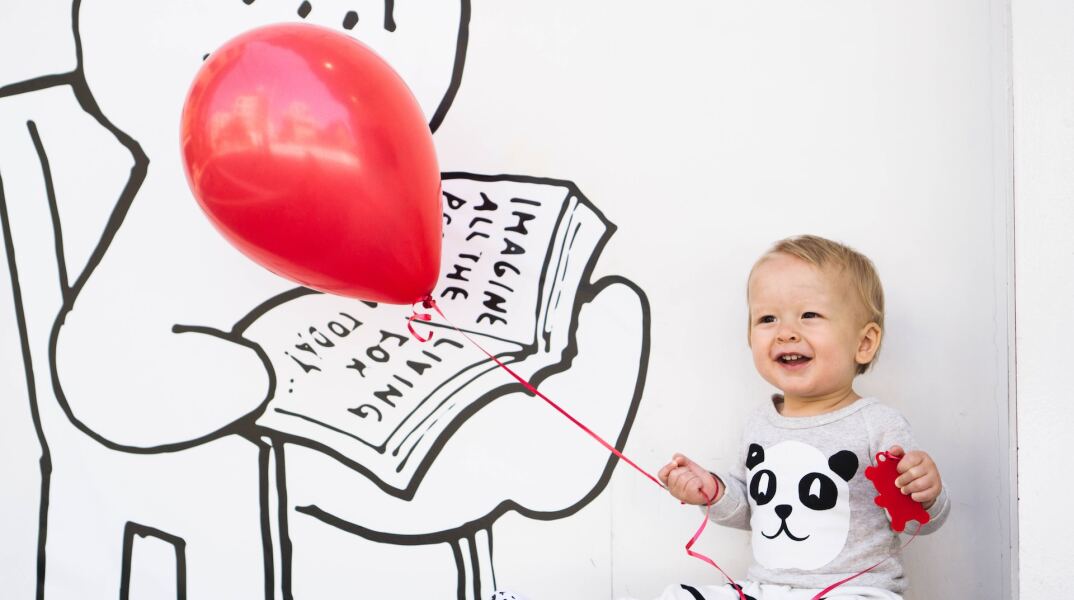 Παιδί κρατά κόκκινο μπαλόνι και ποζάρει σε τοίχο με ζωγραφισμένο βιβλίο