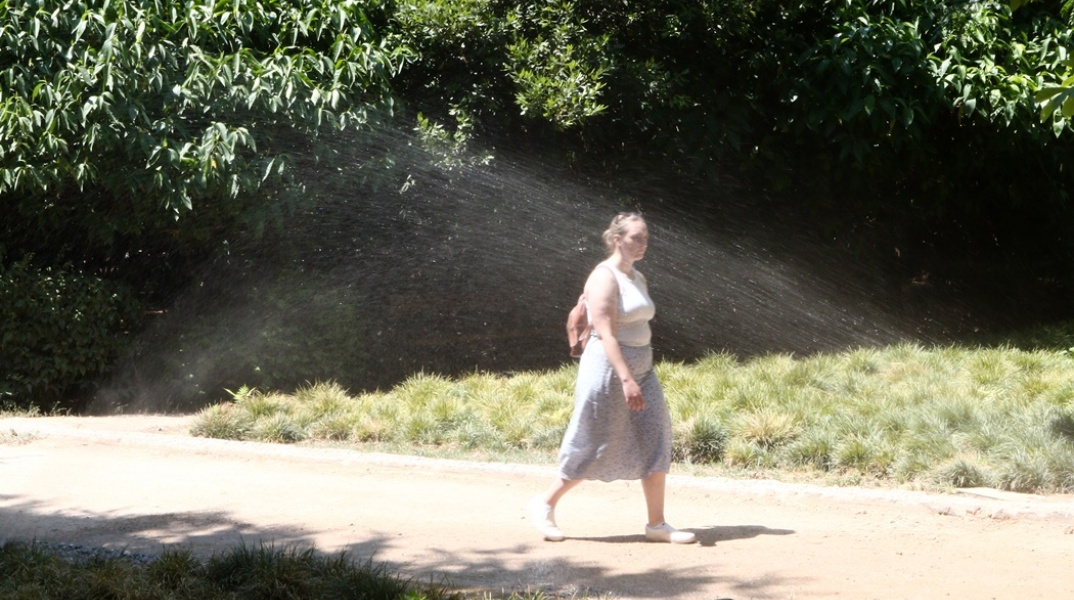 Γυναίκα περπατά σε συνθήκες καύσωνα σε περιβάλλον όπου ποτιστικό δροσίζει φυτά πετώντας νερό