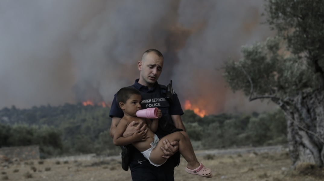 Αστυνομικός μεταφέρει στην αγκαλιά του παιδί ενώ πίσω τους μαίνεται η φωτιά στα Δερβενοχώρια