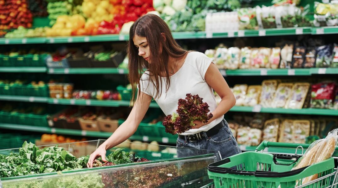 Γυναίκα με το καρότσι του σούπερ μάρκετ επιλέγει λαχανικά από πάγκο