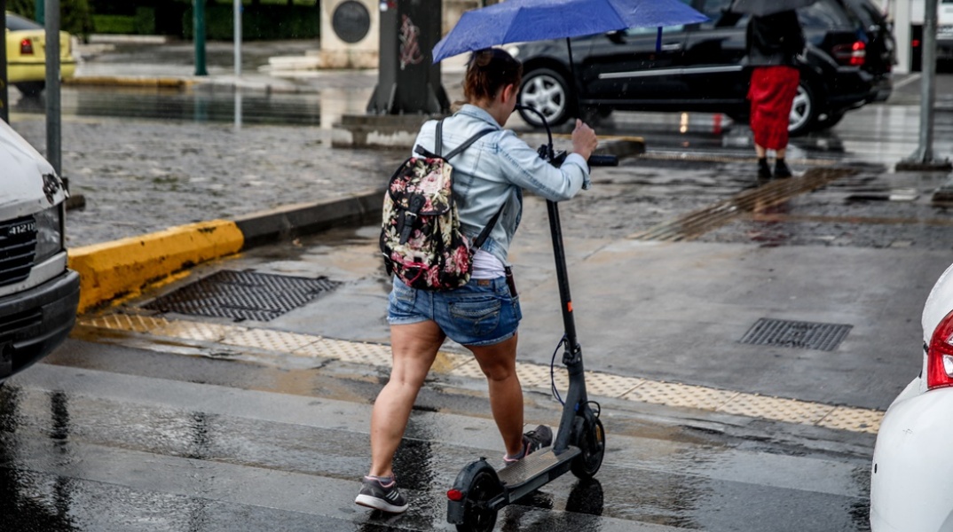 Γυναίκα με πατίνι κρατά ομπρέλα για να προστατευτεί από τη βροχή