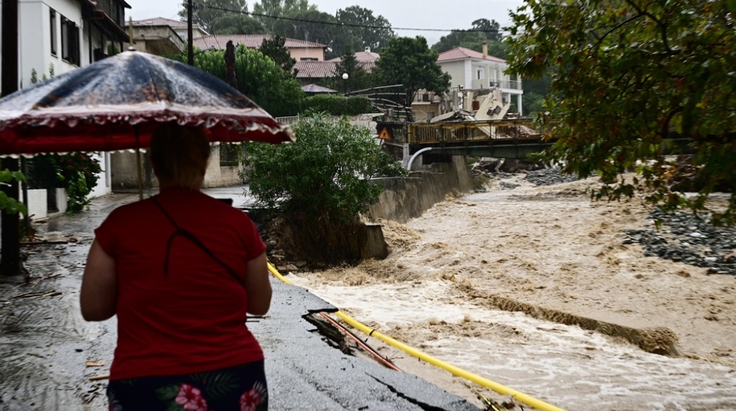 Γυναίκα περπατά με την ομπρέλα της δίπλα σε ποτάμι με ορμητικά νερά