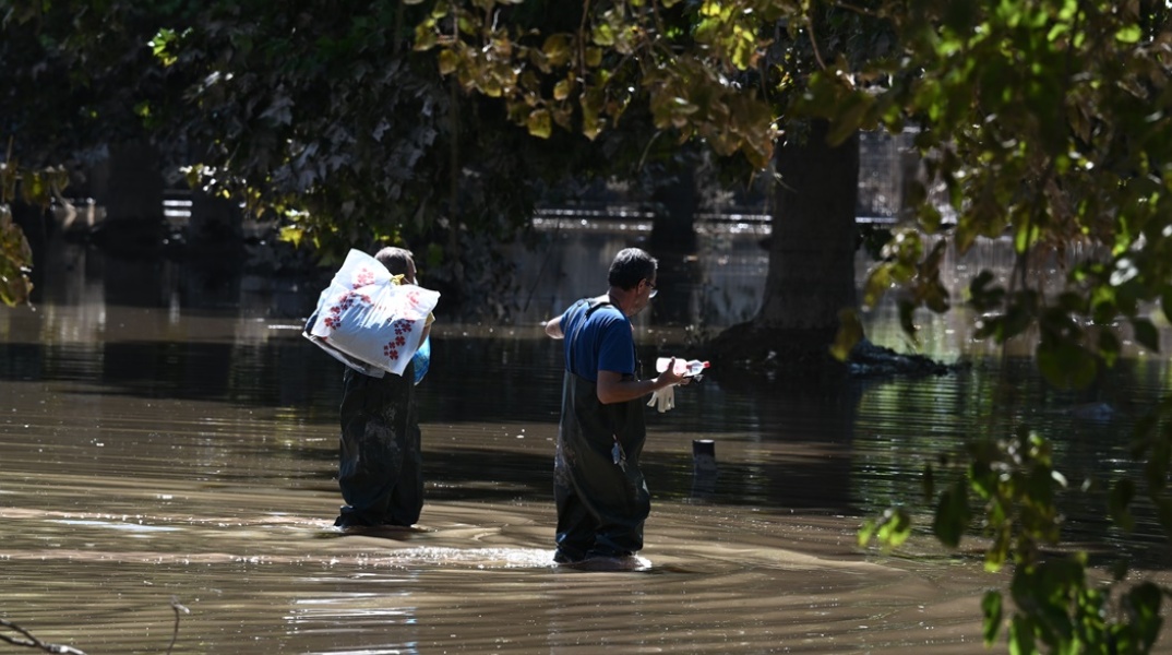 Άνδρες με προστατευτικά περπατούν σε πλημμυρισμένο δρόμο και κουβαλούν πράγματα