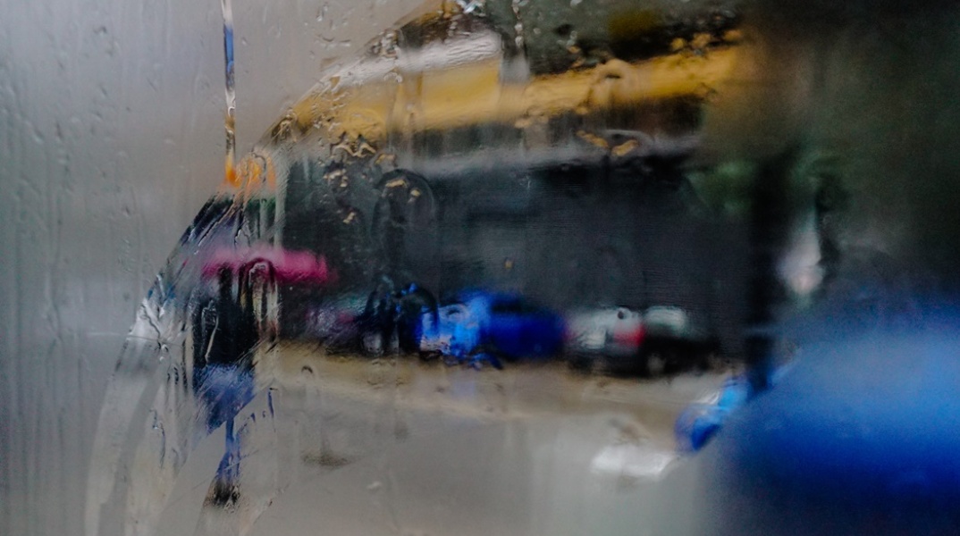 Φωτογραφία μέσα από αυτοκίνητο με θολωμένα τζάμια που δείχνει γυναίκα να κρατά ομπρέλα κοντά σε συρμό του τραμ που διασχίζει τον βροχερό δρόμο