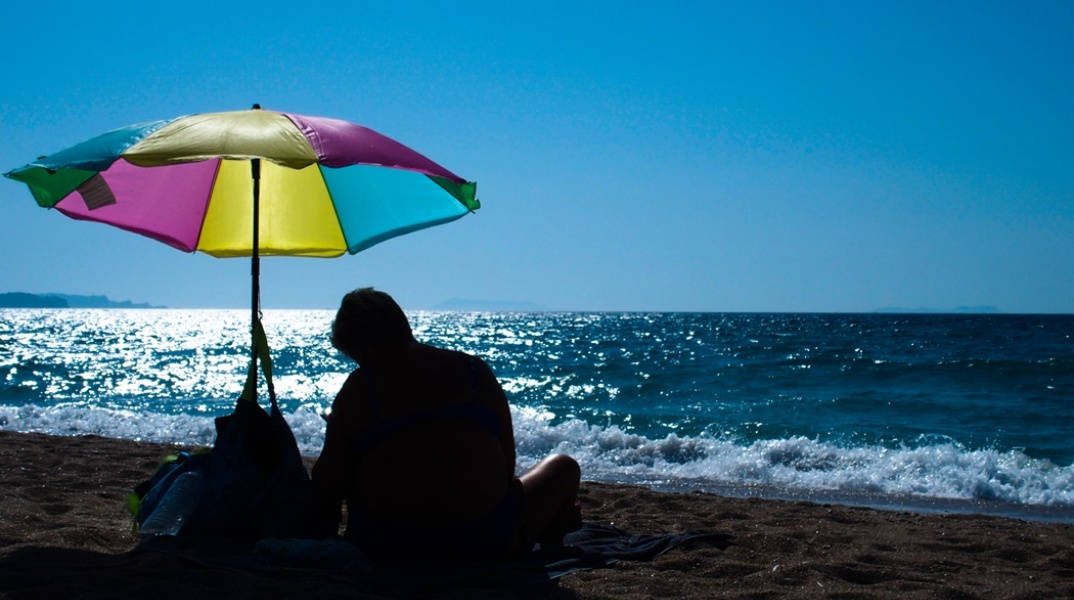 Άνδρας καθισμένος κάτω από πολύχρωμη ομπρέλα θαλάσσης στην ακροθαλασσιά