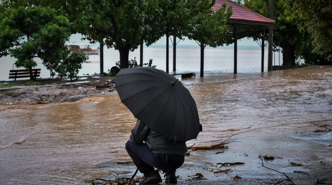 Άνδρας κρατά ομπρέλα για να προστατευτεί από καταιγίδα και στέκεται μπροστά από δρόμο που έχει μετατραπεί σε ποτάμι