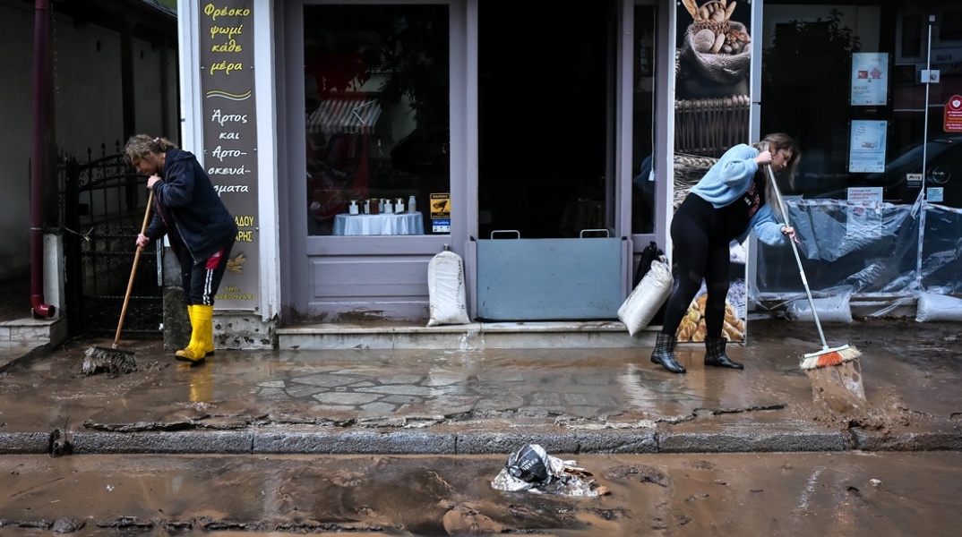 Γυναίκες με γαλότσες και σκούπες ανά χείρας απομακρύνουν από κατάστημα και πεζοδρόμια τα λασπόνερα